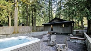 Un jacuzzi, terraza de madera, sillas tipo Adirondack y una cabaña en un área boscosa.