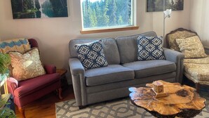 A living room with a grey sofa, patterned cushions, a wooden coffee table, and a window with a view of trees. - Alaskan Adventure Cabin (Sterling)