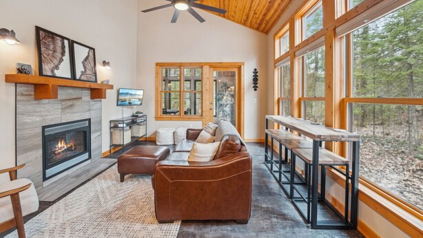 A cozy living room with a fireplace, leather sofa, and large windows offering a forest view.