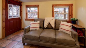 A cozy room with a brown leather sofa, two windows, a wooden door, and a small table with a plant.