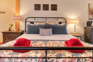 A neatly made bed with floral bedding, two red towels, and a striped pillow. There are two bedside tables with lamps, a small clock, and a vase with flowers. The room has a wooden headboard and a painting on the wall. - The Willow Cottage (Cali Cochitta ) (Moab)
