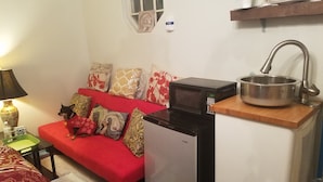 A compact kitchenette with a microwave, a sink with a pot, and a red sofa with decorative pillows. - Tinyhouse, Enchanted Cottage nestled in the backyard of the fenced in property. (Atlanta)