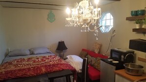 A bedroom with a large bed, a chandelier, a small table with a lamp, a television, and a kitchenette area. - Tinyhouse, Enchanted Cottage nestled in the backyard of the fenced in property. (Atlanta)