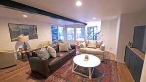 Een moderne woonkamer met een bruine leren bank, beige fauteuils, een witte salontafel en een groot vloerkleed. Er zijn ramen met uitzicht op bomen, een schilderij aan de muur en een kast met spiegel.