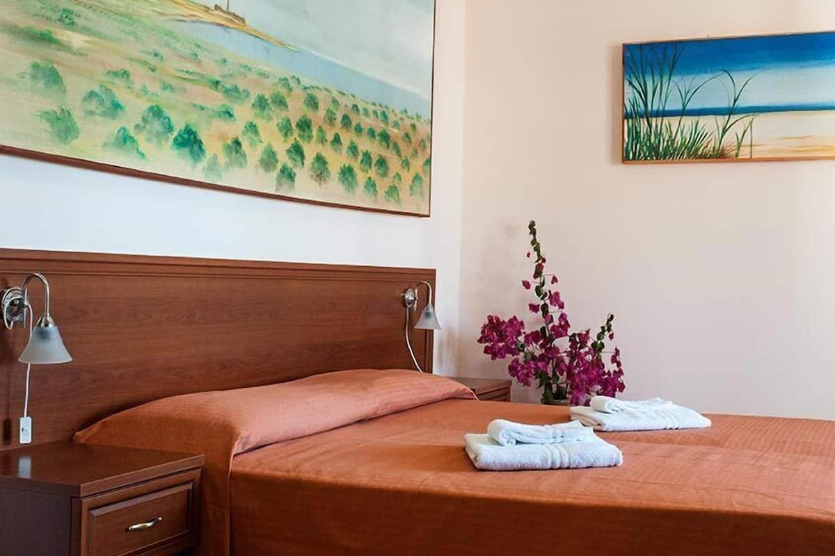 Quarto de hotel com cama, mesa de cabeceira, luminária, pintura de paisagem e um vaso com flores.