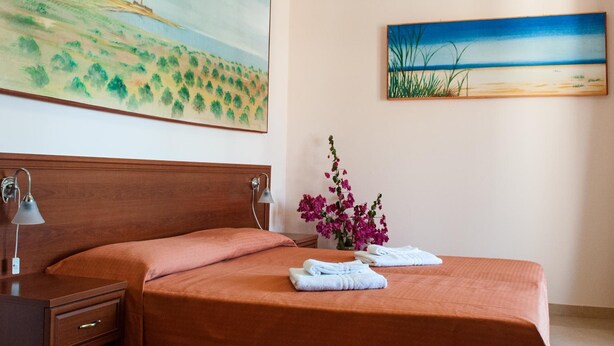 Uma cama bem arrumada com um espalha cama marrom, um abajur, um vaso com flores cor-de-rosa e duas toalhas dobradas.