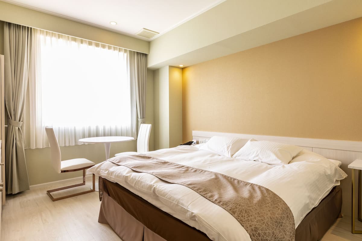 A hotel room with a bed, a chair, a small table, and a window with curtains.