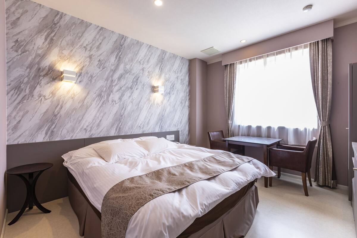 A modern hotel room with a marble wall, a bed with white linens, a desk, and a chair.
