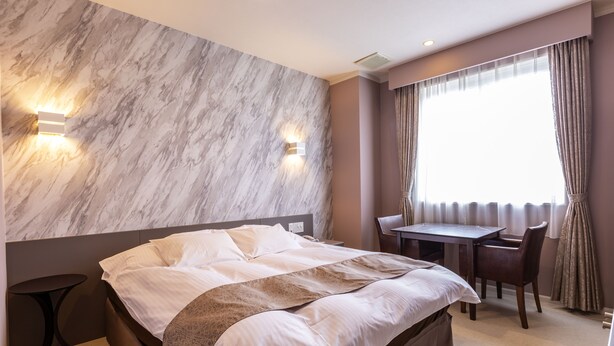 Een hotelkamer met een bed, een bureau en een stoel. Twee wandlampen en een raam met gordijnen.