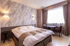 Une chambre d’hôtel avec un lit, un bureau et une chaise. Il y a deux lampes fixées au mur et une fenêtre avec des rideaux.