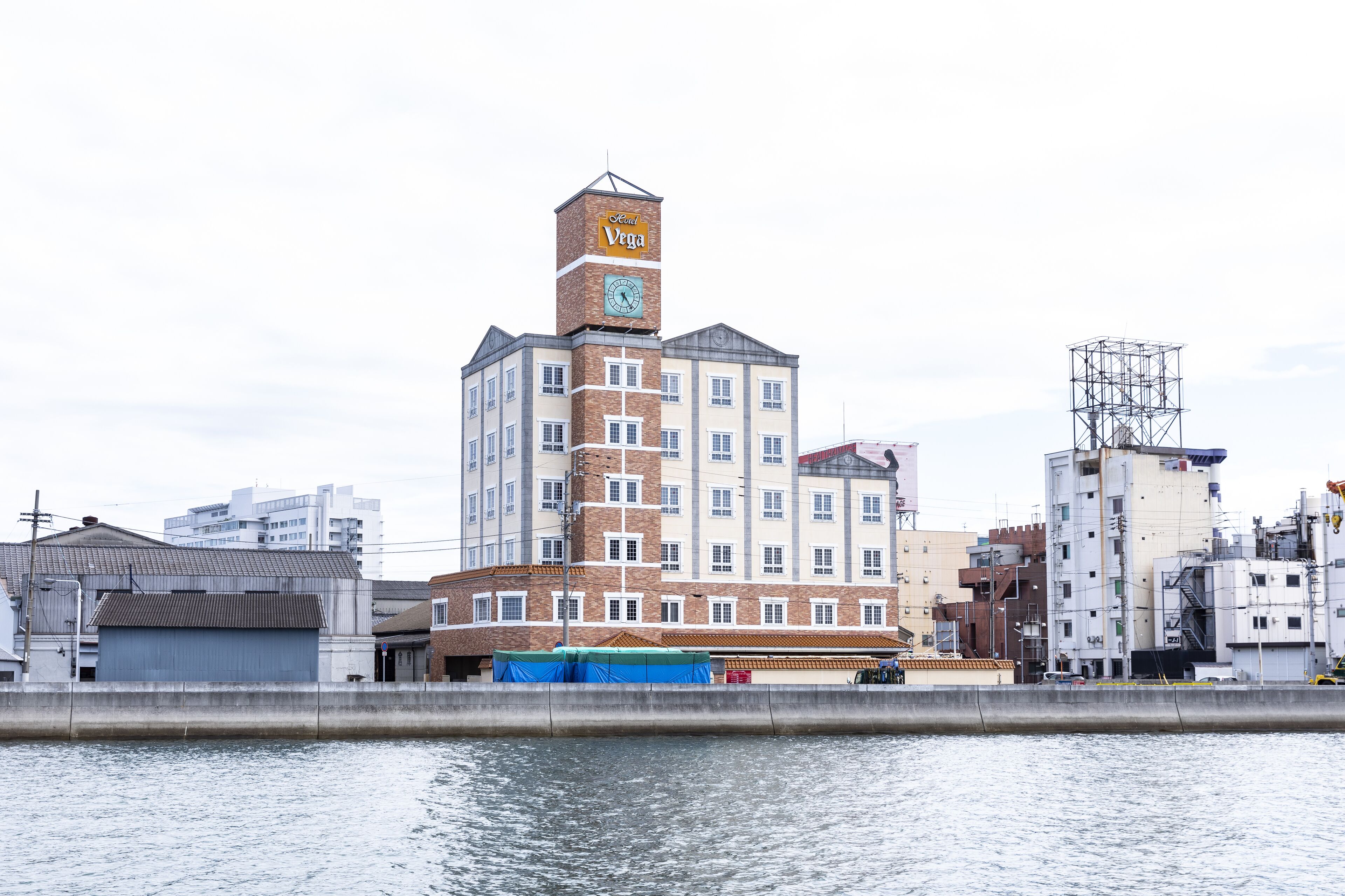 Un edificio de varios pisos con una torre de reloj, un área techada con lona azul y vista al agua.