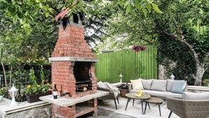 A brick outdoor kitchen with a chimney, surrounded by wicker furniture and a green fence. - Holiday home with heated outdoor swimming pool up to 12 persons (Cluj-Napoca)
