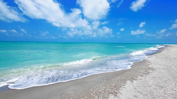 Una playa con aguas turquesas cristalinas y una orilla arenosa bajo un cielo azul brillante con nubes dispersas.