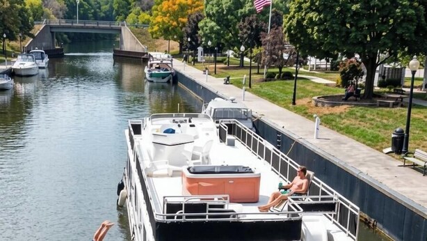 Una casa flotante atracada en un canal, con una persona sumergiéndose en el agua, un pequeño muelle y un puente al fondo.