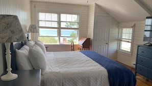 A bedroom with a bed, a nightstand, a lamp, a dresser, and a view of the ocean through large windows. - Quintessential New England Home on Private Beach  (Westerly)
