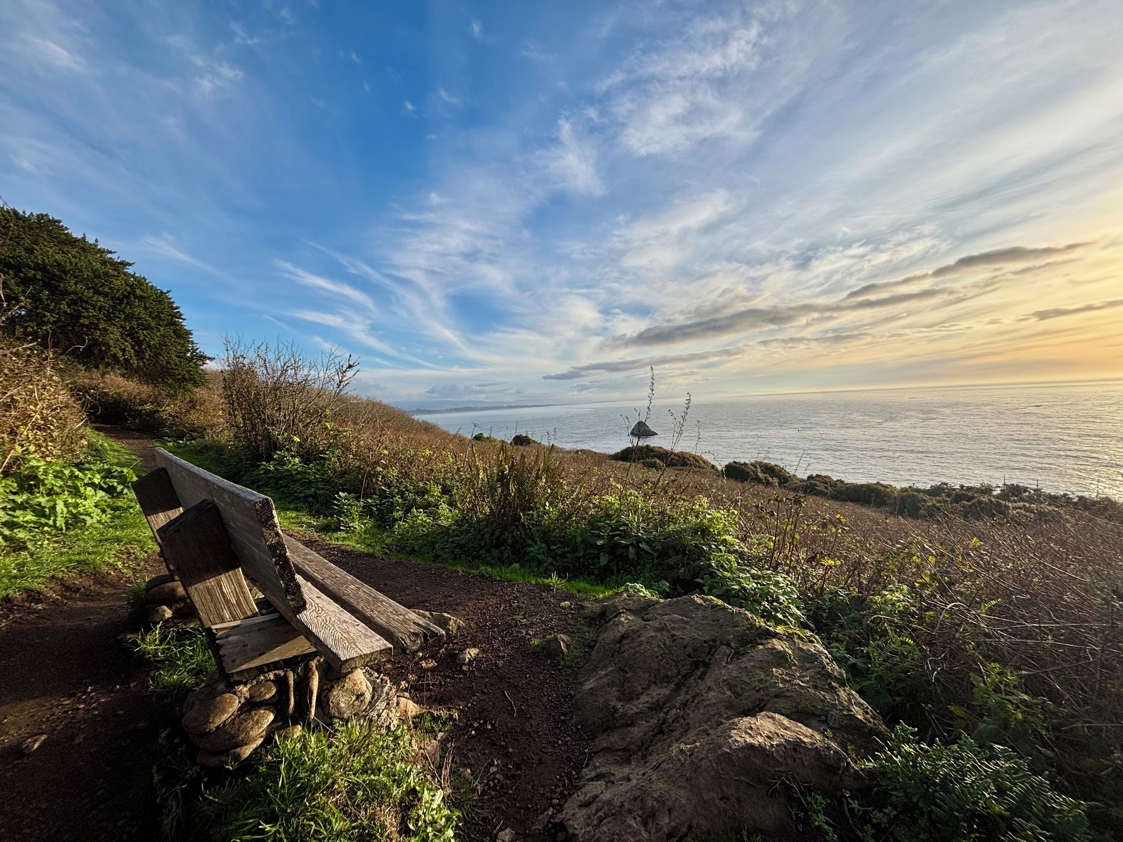 A wooden bench on a rocky path overlooking a coastal area with a view of the sea and a clear sky.