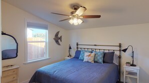 A bedroom with a bed, a ceiling fan, a window, a nightstand, and a wall-mounted mirror.