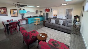 Una sala de estar moderna con un sofá gris, sillones rojos con estampado floral, una mesita redonda y una zona de cocina con armarios verde azulado y un ventilador de techo.