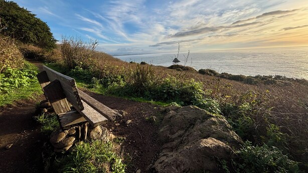 Un banco de madera en un sendero rocoso con vistas a la costa, el mar y un cielo despejado.