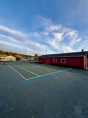 An outdoor tennis court with a net, surrounded by a red building and a fence.