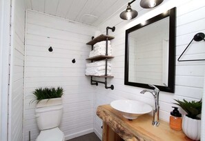Ein Badezimmer mit einer weißen Toilette, einem hölzernen Waschtisch mit Spüle, einem Spiegel und einem Ablagebereich mit Handtüchern.