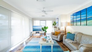 A spacious living room with a large TV, a ceiling fan, and a view of the sea through the windows. - Bay Parklands, 49 2 Gowrie Ave - ducted aircon, Wifi, views, pool, tennis (Nelson Bay)