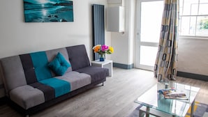 A modern living room with a grey and blue sofa, a glass coffee table, a white side table with a vase of flowers, and a large window with curtains. - Westeria Cottage (Barnstaple)