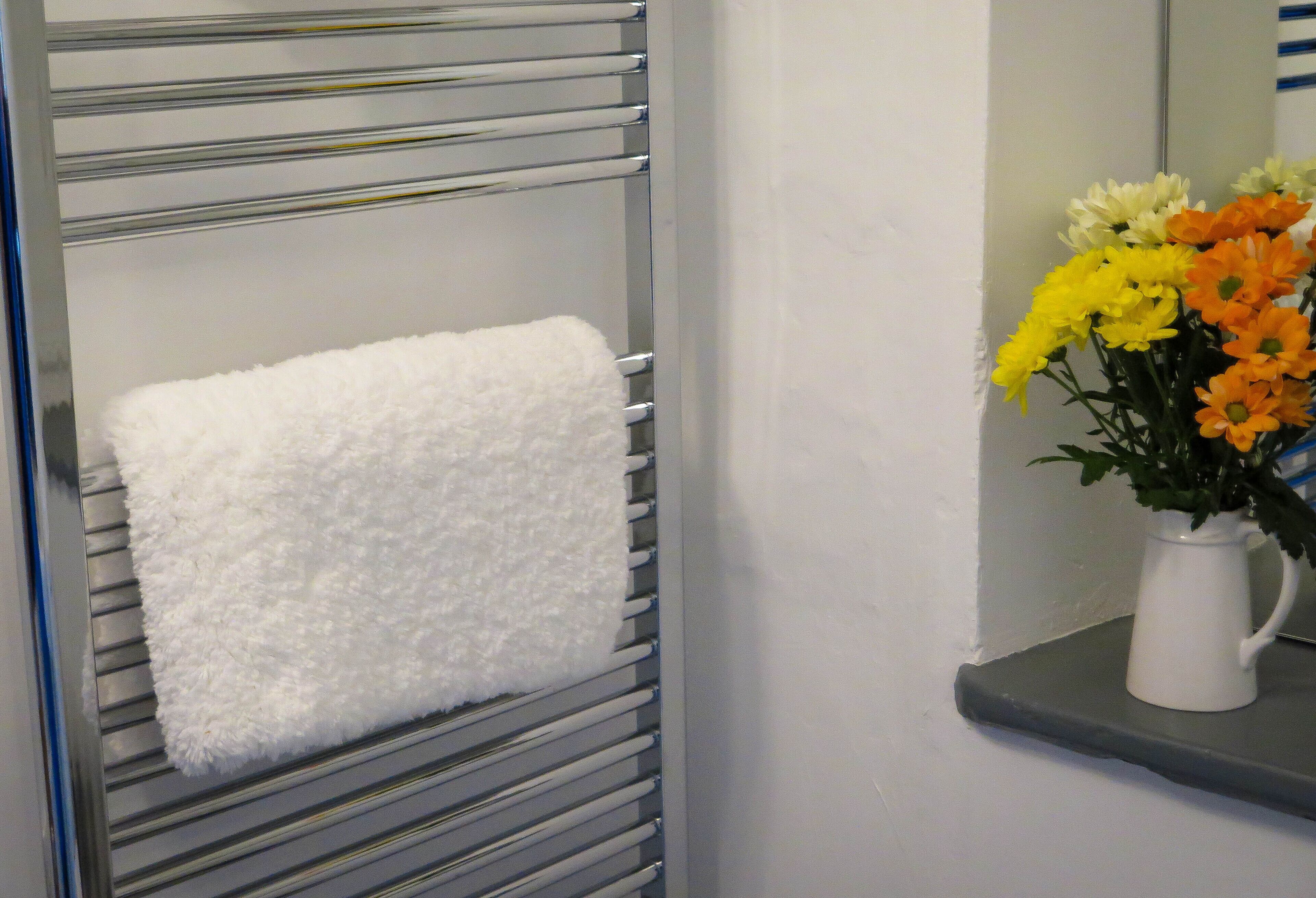 A bathroom with a towel rack holding a white towel, a white vase with flowers, and a mirror on the wall.