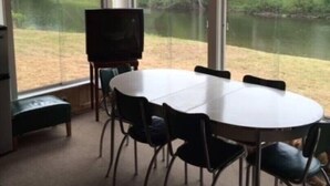 Une pièce avec une table ronde et des chaises, un téléviseur et une vue sur un lac, visible à travers de grandes fenêtres.