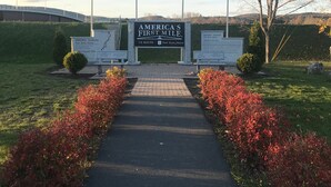 Une signalisation marquant le début du premier kilomètre d’une route américaine, avec plusieurs drapeaux, un chemin pavé et une végétation luxuriante tout autour.