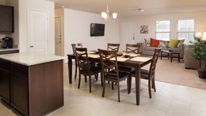 Una cocina moderna con una isla central, mesa de comedor y sillas. Hay una sala de estar con un sofá y un televisor montado en la pared.