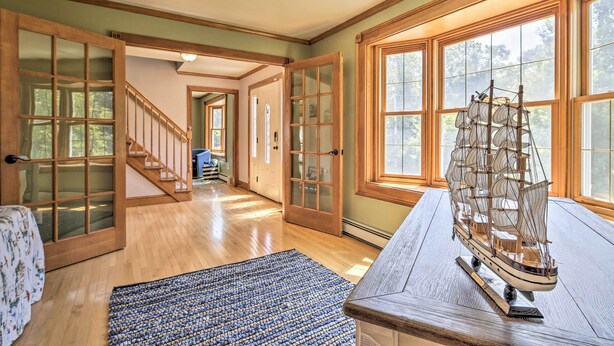 A spacious room with a wooden staircase, large windows, and a model ship on a table. - Walk to Buzzards Bay Beach: Spacious Onset Home (Onset)