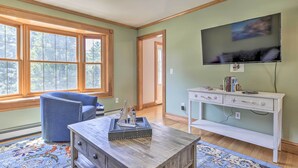 A room with a wooden coffee table, a blue armchair, a white console table with drawers, a flat-screen TV mounted on the wall, and a large window with a view of trees. - Walk to Buzzards Bay Beach: Spacious Onset Home (Onset)