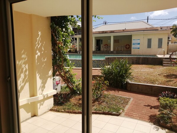 On peut voir une piscine, une terrasse carrelée, un petit jardin avec des plantes et un bâtiment dont le toit est recouvert de tuiles.