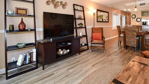 A living room with a TV, shelving, a chair, and a dining area. - Aldea A8 Beachfront 2 Bedroom 2 Bath - Tranquil Belleair Beach (Belleair Beach)