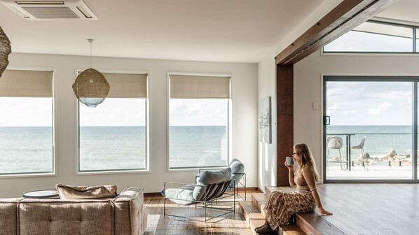 A spacious room with large windows overlooking the sea, a modern chair, and a sofa.