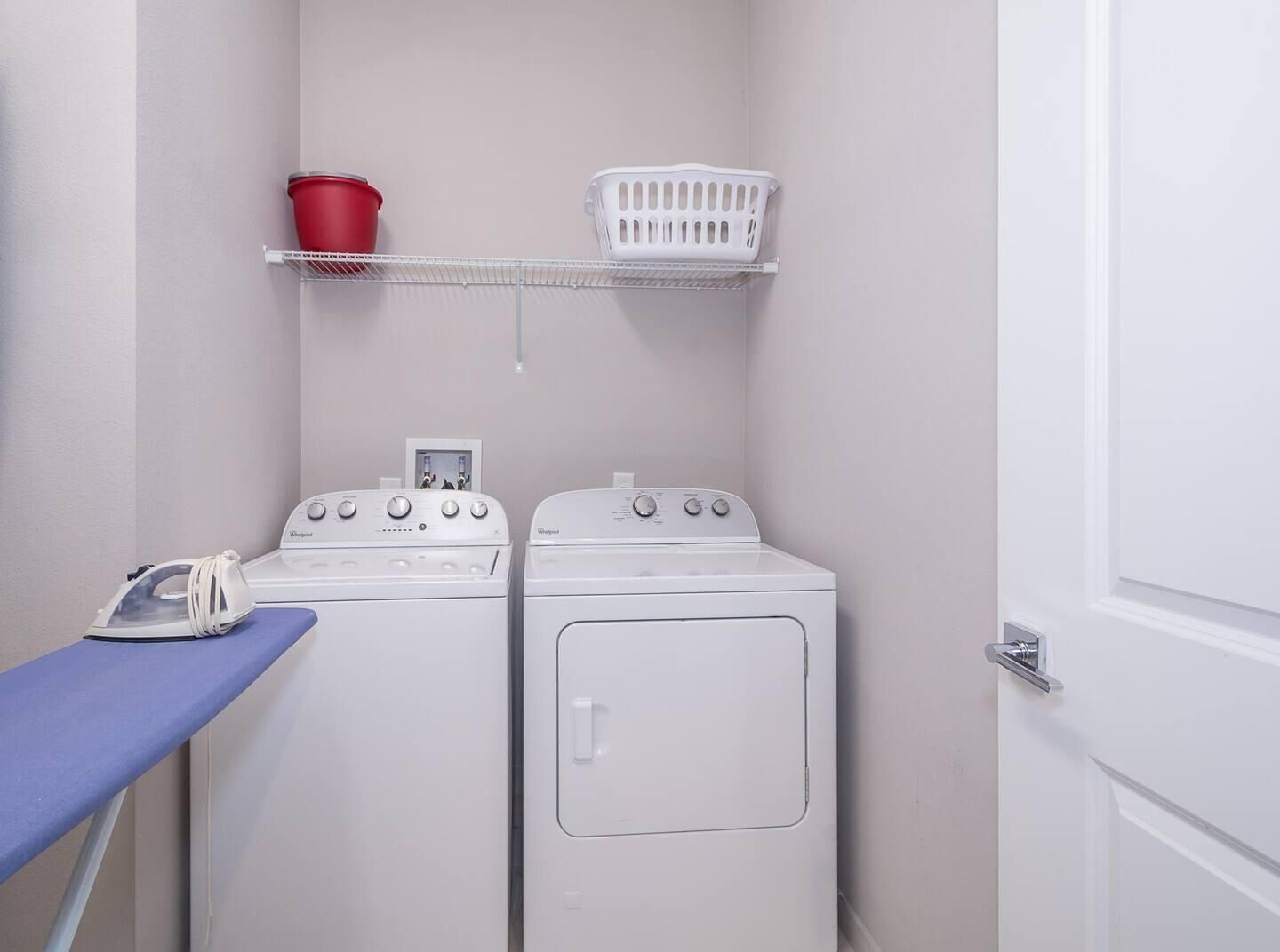 Une buanderie équipée de deux machines à laver et de deux sèche-linge blancs, d’un seau rouge, d’un panier à linge blanc et d’une planche à repasser bleue.