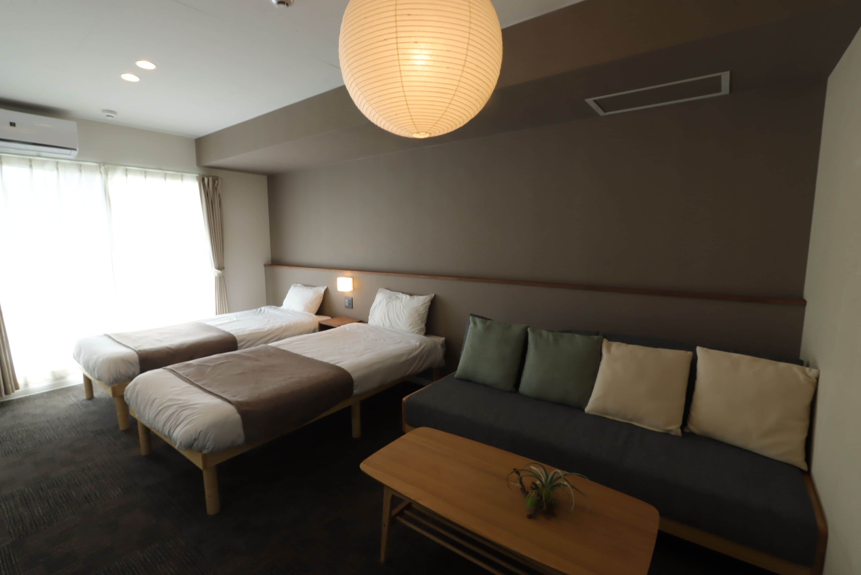 A hotel room with two single beds, a sofa, a wooden coffee table, and a hanging paper lantern.