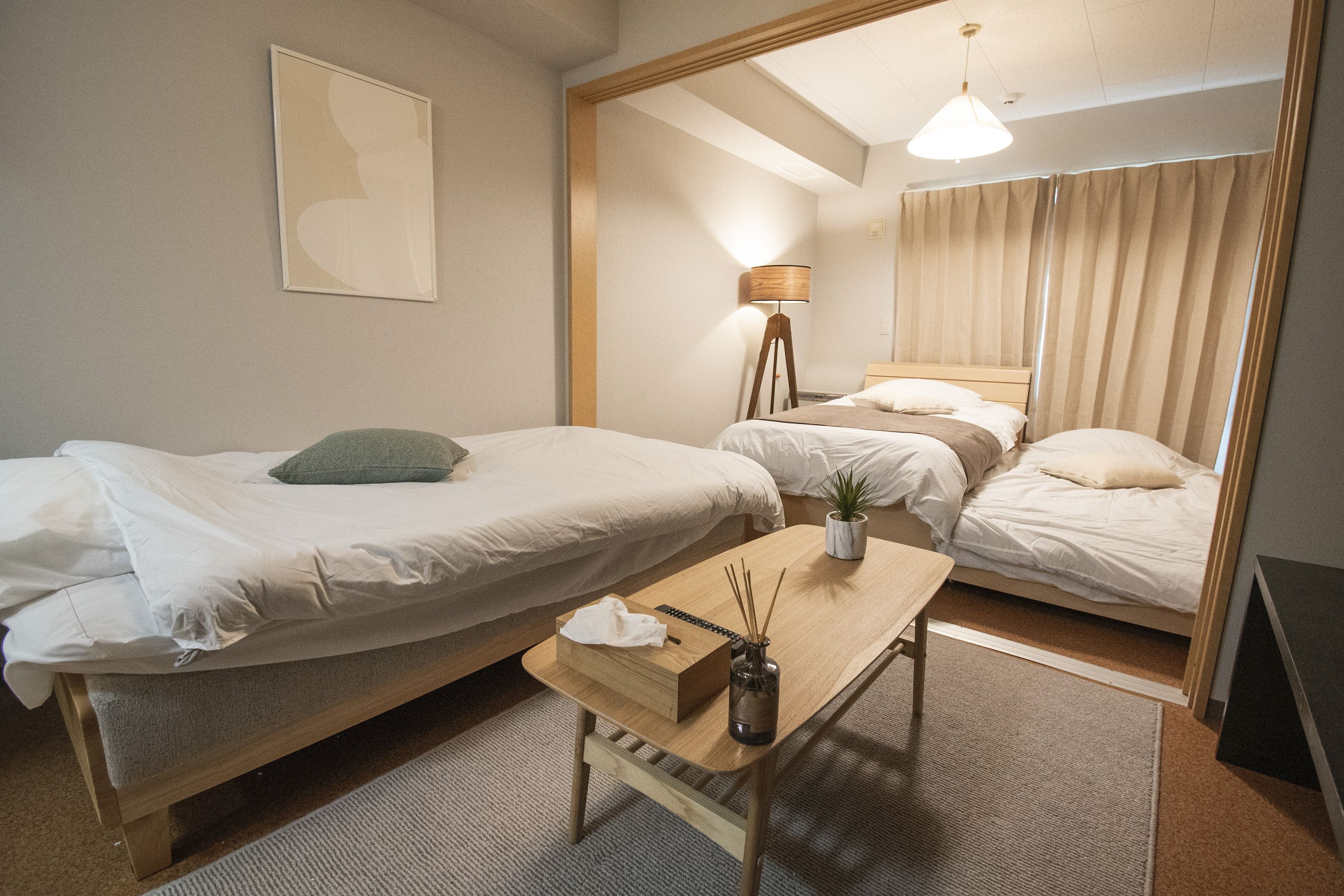 A hotel room with two single beds, a small table with a vase and a box, a standing lamp, and a large mirror.