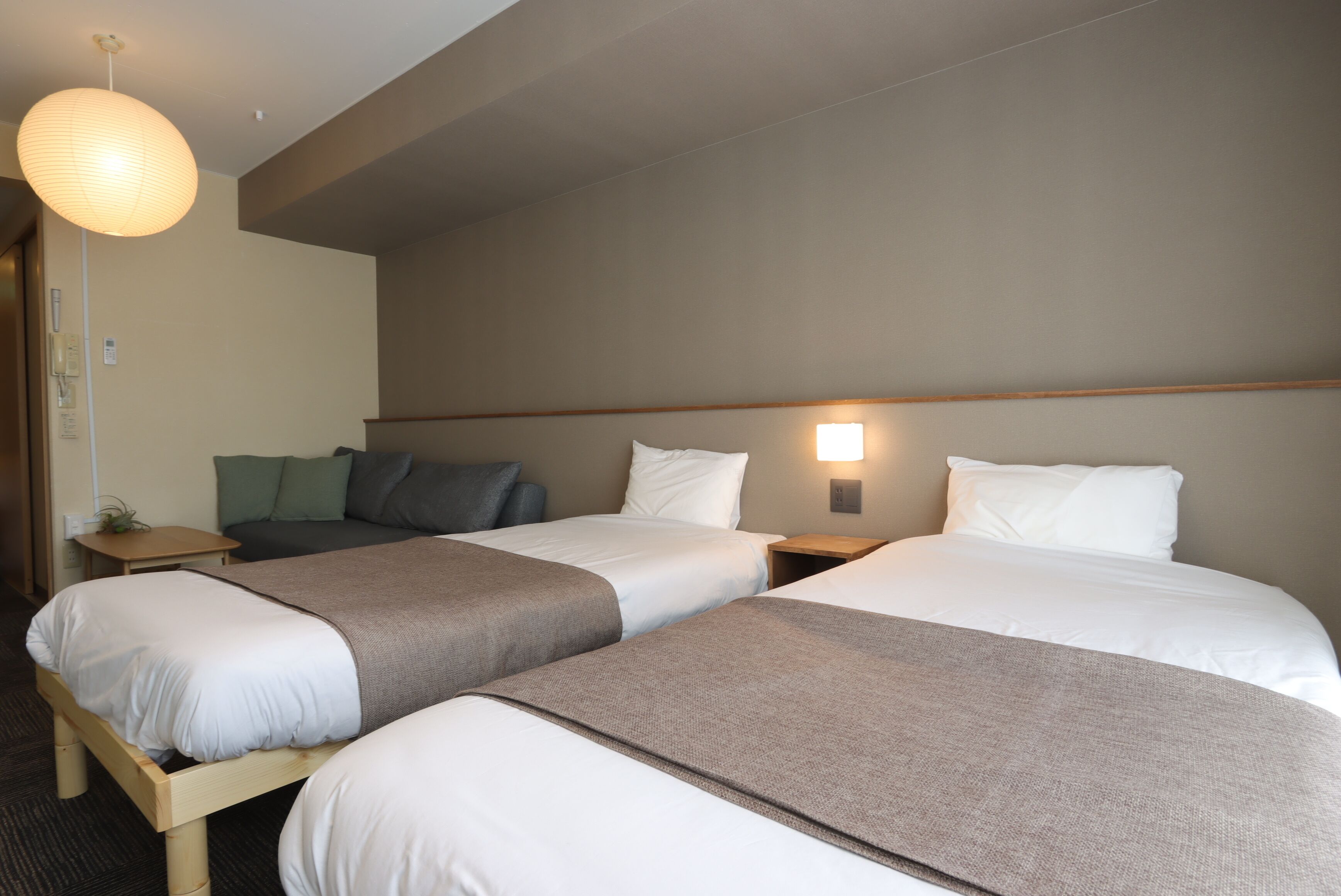 A hotel room with two beds, a bedside table, a lamp, and a wall-mounted light.