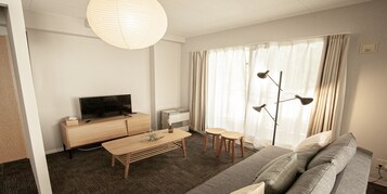 Habitación de hotel moderna con un mueble bajo de televisión de madera, un televisor de pantalla plana, una mesita de centro de madera, un sofá gris y un ventanal con cortinas translúcidas.
