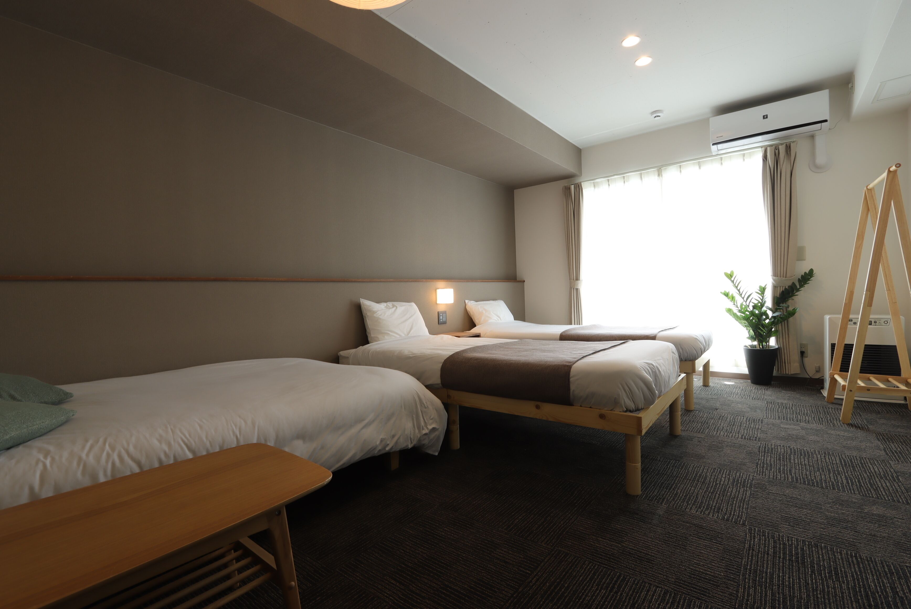 Une chambre d’hôtel avec deux lits simples, un banc en bois, une plante en pot et une échelle appuyée contre le mur.