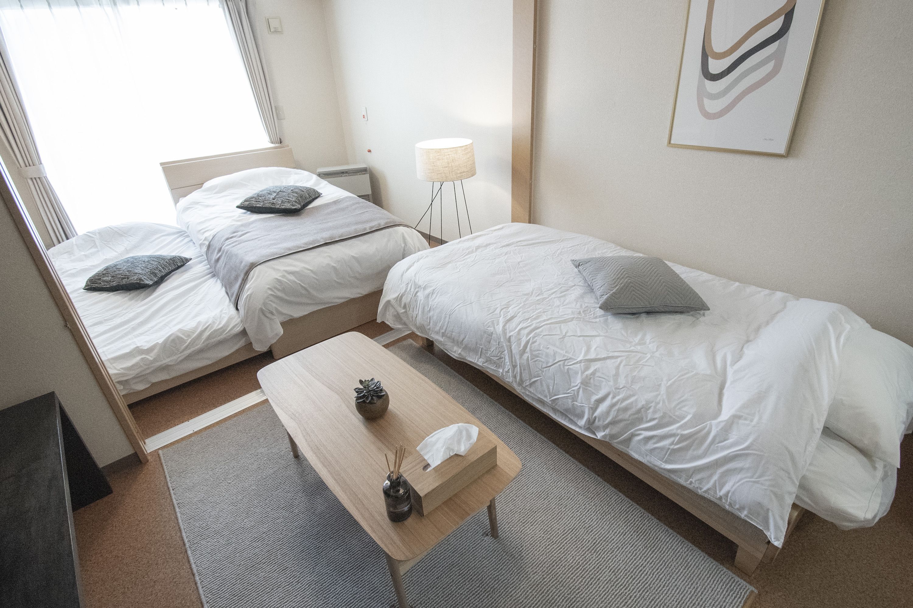 Ett hotellrum med två separata sängar, ett litet träbord, en lampa och ett inramat, abstrakt konstverk på väggen.