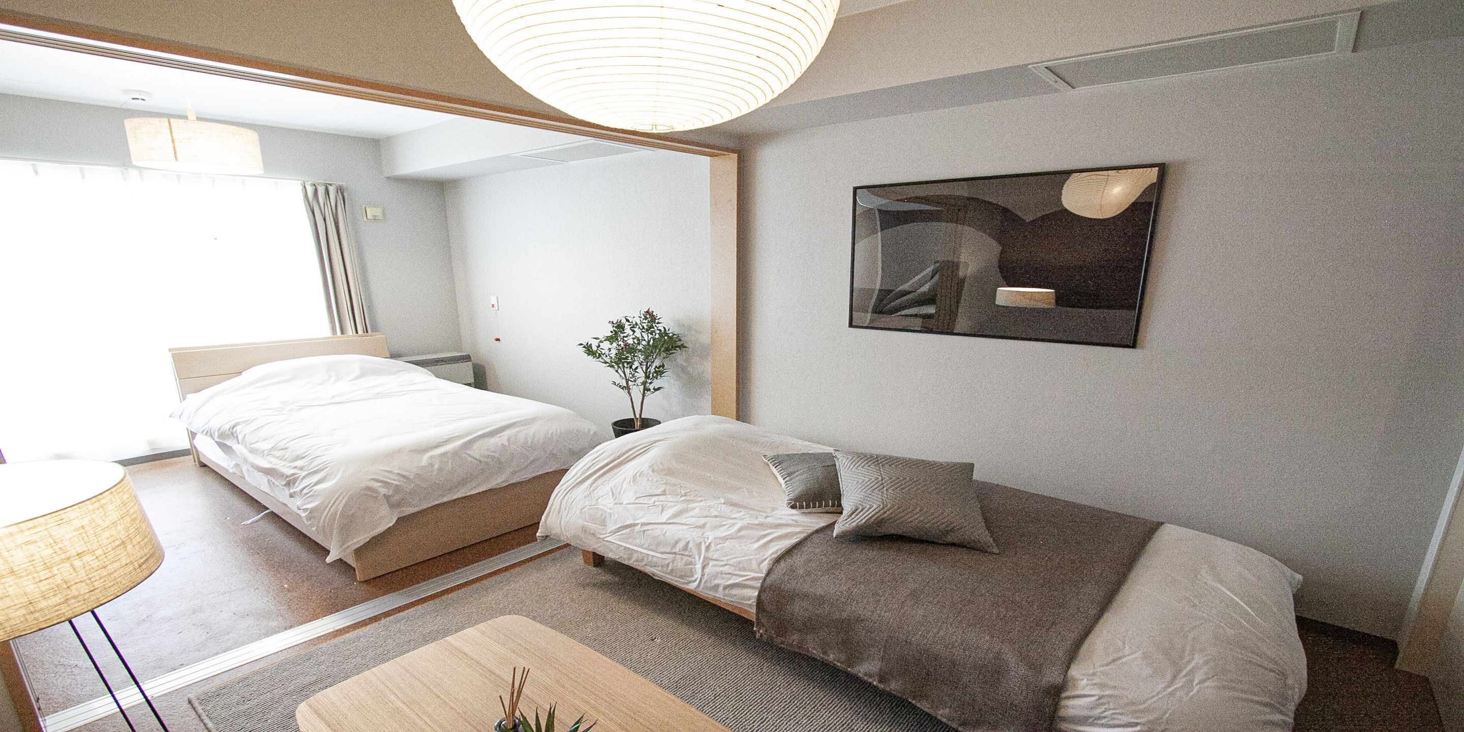 A modern hotel room with two beds, a small table, a potted plant, and a large abstract painting on the wall.