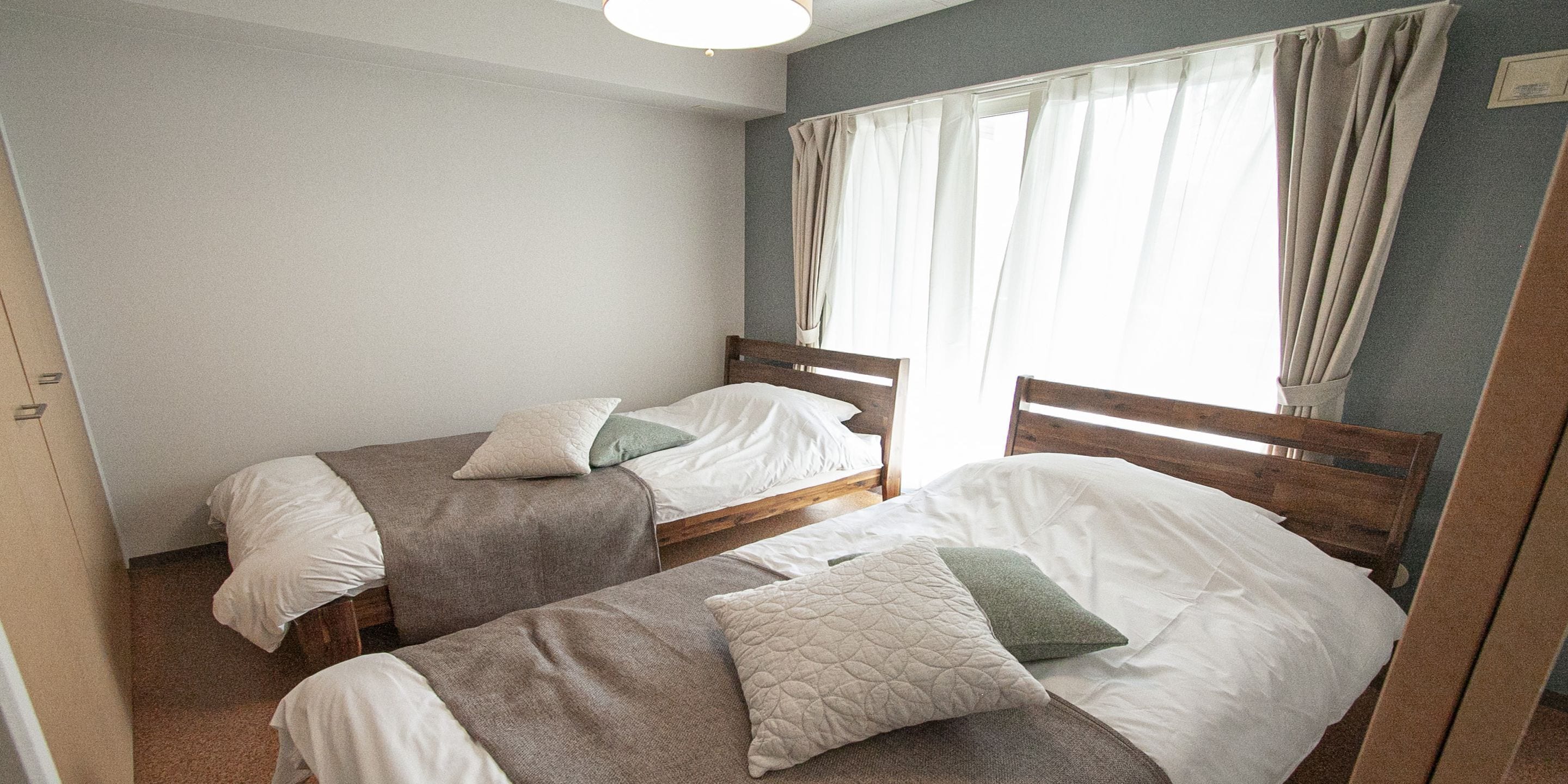 A room with two single beds, a window with curtains, and a plain wall.