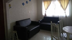 Un salón con dos sofás negros, una mesita redonda y una silla. Hay dos ventanas con cortinas traslúcidas.