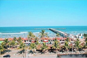 A beachside view with a pier extending into the ocean, palm trees, and beach umbrellas.