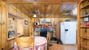 Interior de una cabaña de madera con una mesa de comedor lista, sillas, una chimenea de piedra y un refrigerador blanco.