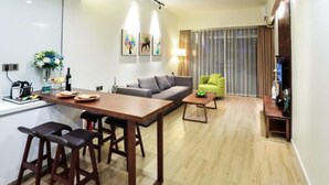 Un salon moderne comprenant un canapé, un fauteuil vert, une table à manger en bois avec des tabourets de bar, et un coin cuisine avec un comptoir et des appareils électroménagers.