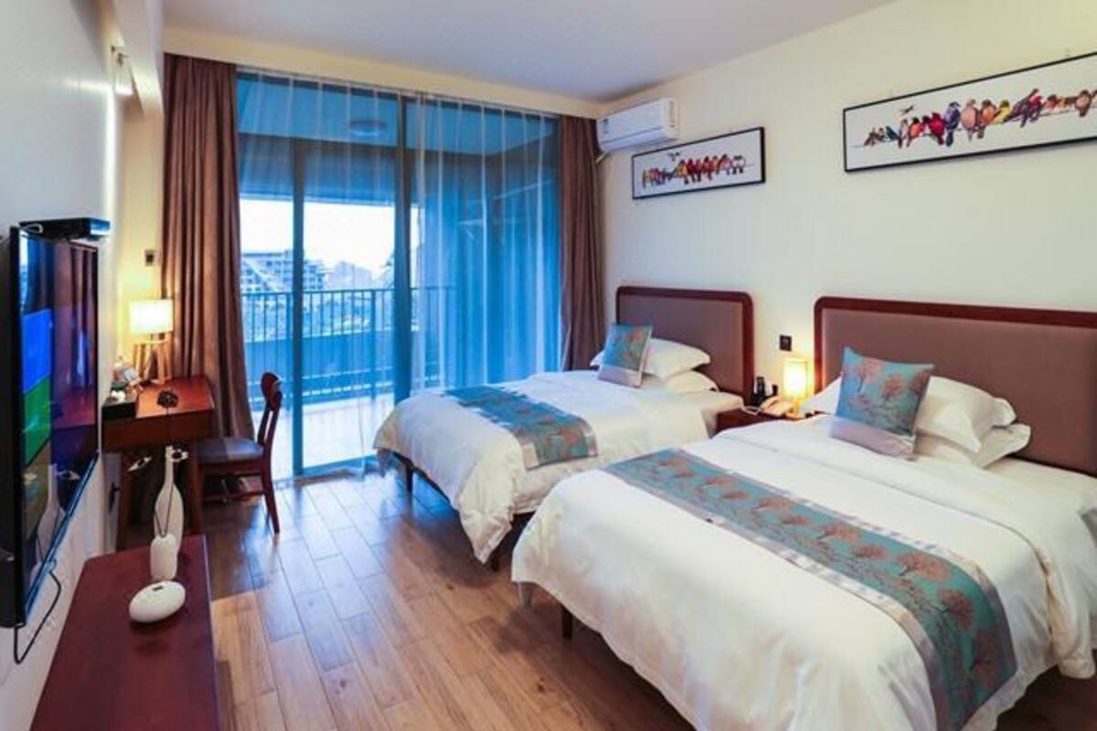 Ett hotellrum med två sängar, ett skrivbord, en stol, en TV och en balkong med gardiner.